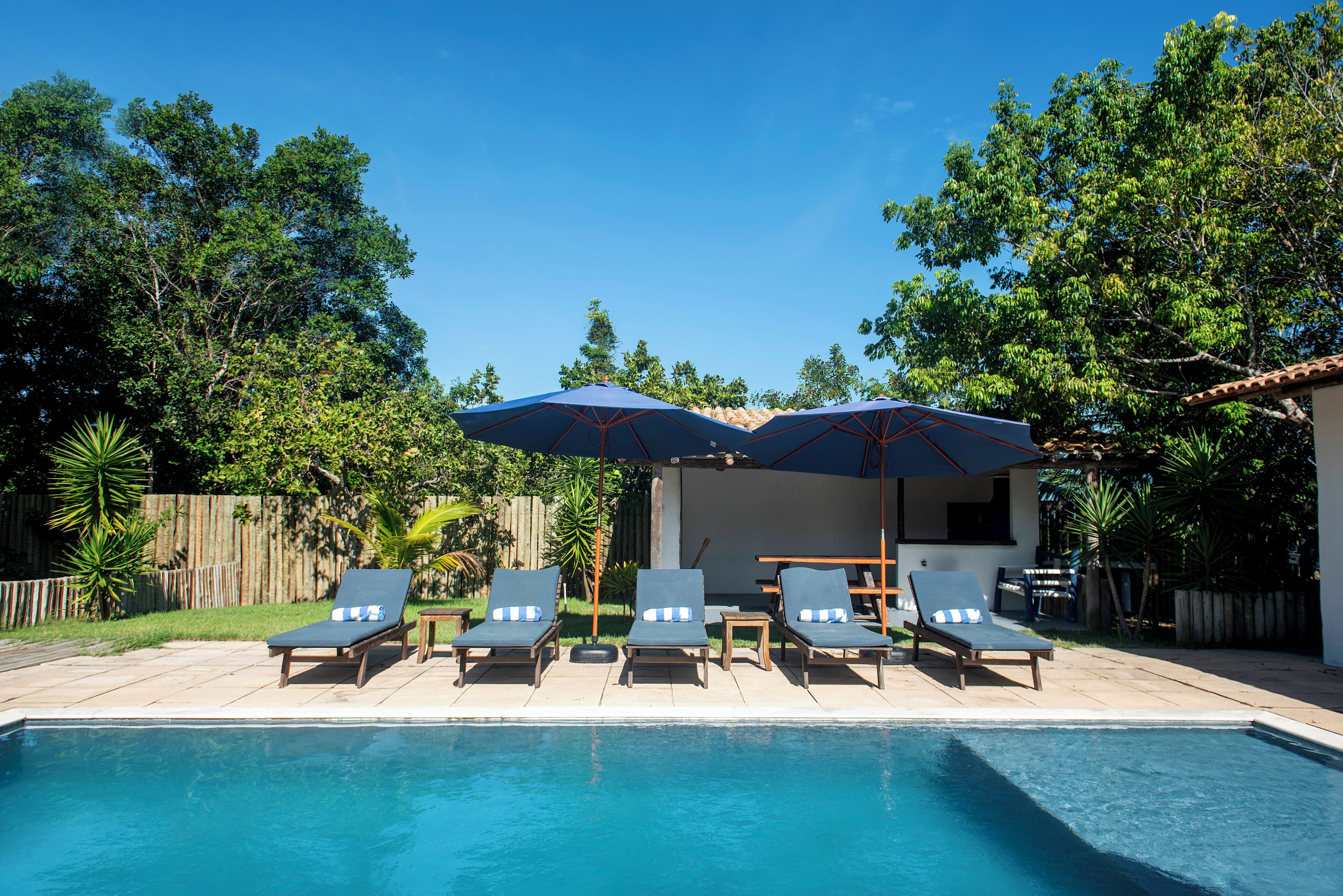 Une piscine entourée d’arbres et d’une clôture en bois, avec des chaises longues et des parasols à proximité.