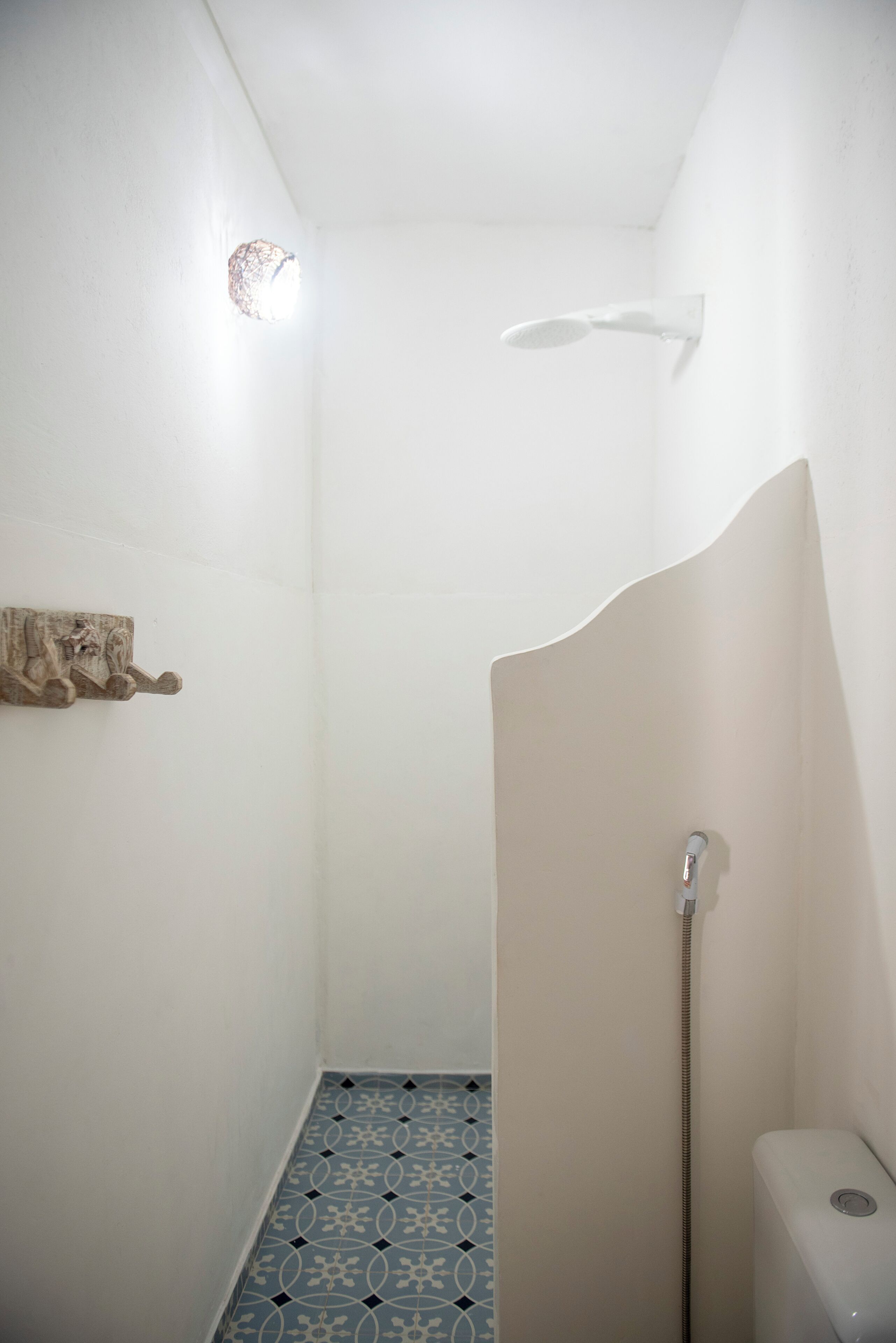 A small, narrow shower with a white wall, a blue patterned floor, and a wooden shelf.