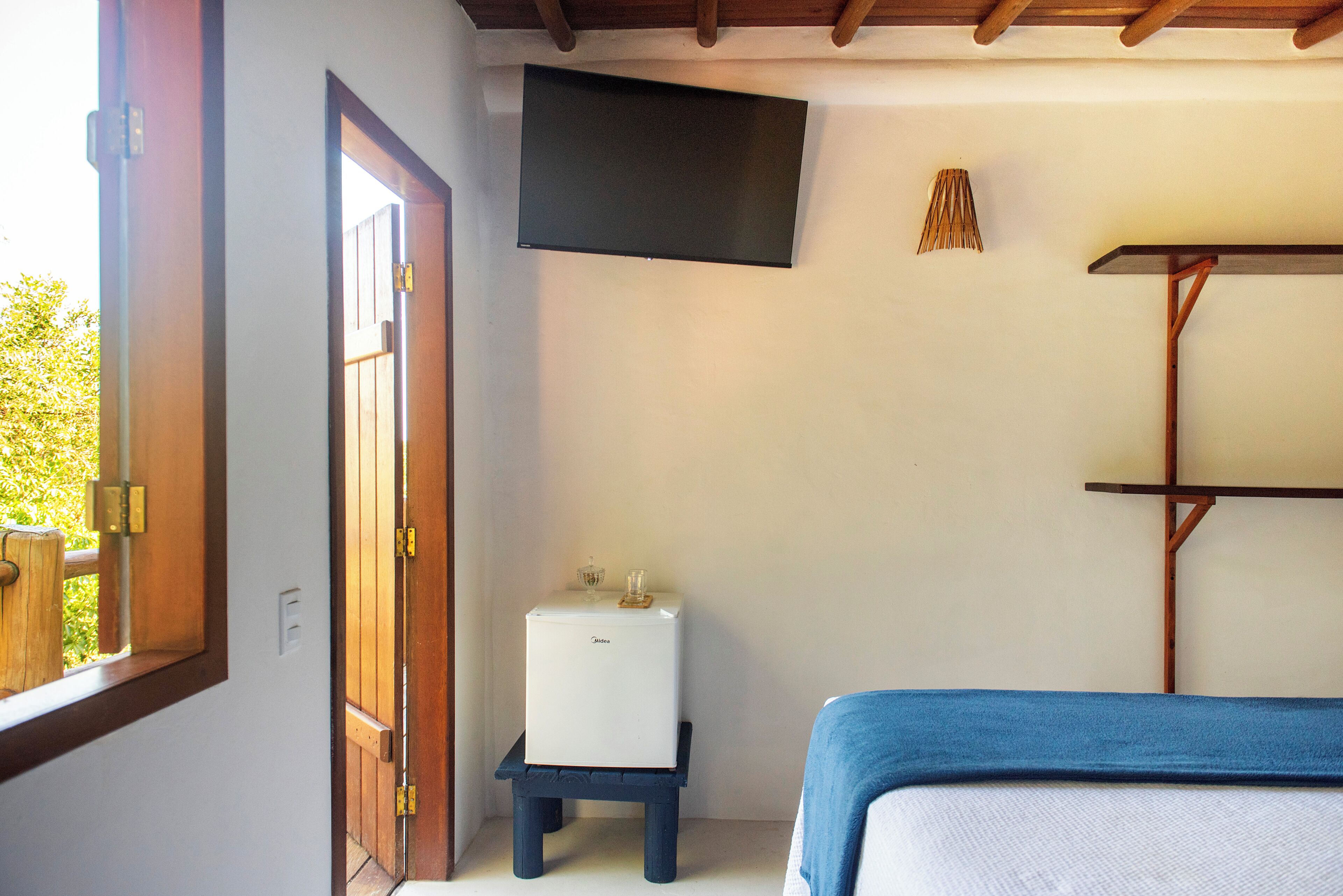 A room with a bed, a small white appliance on a stand, a mounted flat-screen TV, and a wooden door.