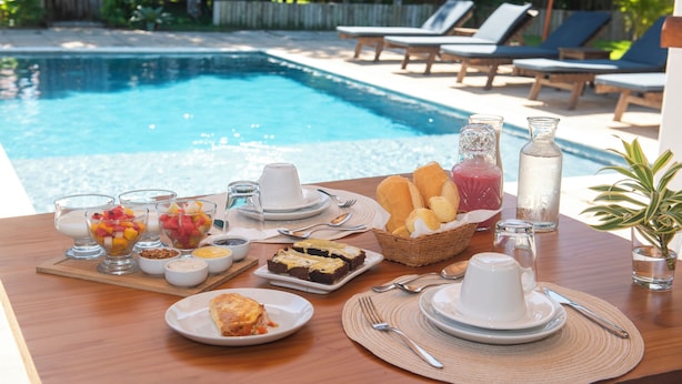 A poolside breakfast setup with various dishes, a bottle of water, and lounge chairs in the background. - Pousada Lua Cheia Praia do Espelho (Porto Seguro)