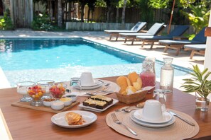 A poolside breakfast setup with various dishes, a bottle of water, and lounge chairs in the background. - Pousada Lua Cheia Praia do Espelho (Porto Seguro)