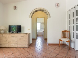 A room with a TV on a wooden cabinet, a chair, a tiled floor, and a bathroom visible through an archway. - Malù House by Interhome (Villasimius)