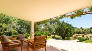 Una veranda riparata con mobili in legno, un pilastro in pietra e vista su un giardino rigoglioso e delle colline.