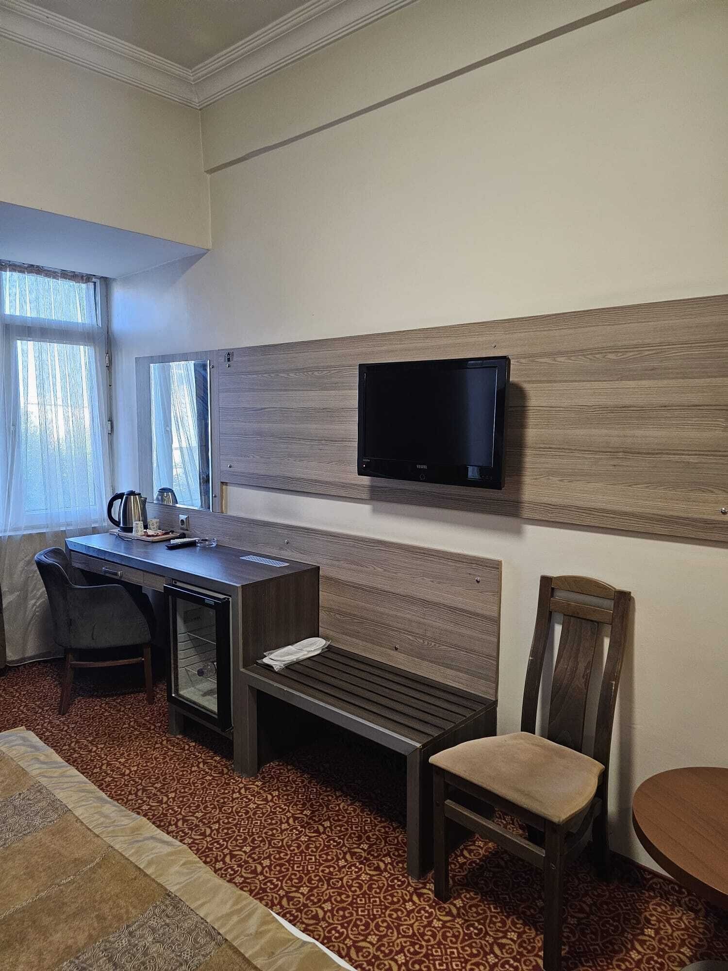 A hotel room with a bed, a desk, a chair, a television mounted on the wall, and a window with curtains.