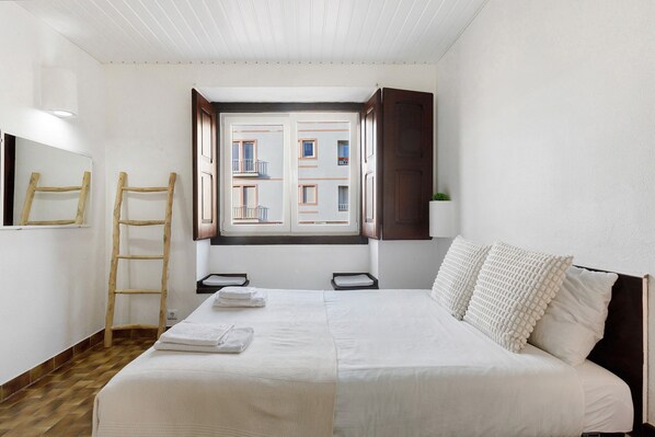 Ein Schlafzimmer mit einem großen Bett, einer Holzleiter, einem Fenster mit Blick auf Gebäude und einer kleinen Pflanze auf dem Nachttisch.