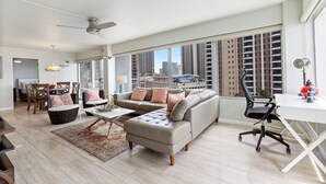 A spacious living room with a large sofa, a dining area, and a view of tall buildings through large windows. - Ilikai Hotel Condo with Ocean, Lagoon & Fireworks Views (Honolulu)