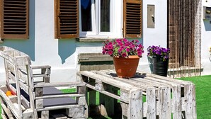 Un banc et une table en bois, avec des pots de fleurs devant un bâtiment aux volets en bois et doté d’une fenêtre.