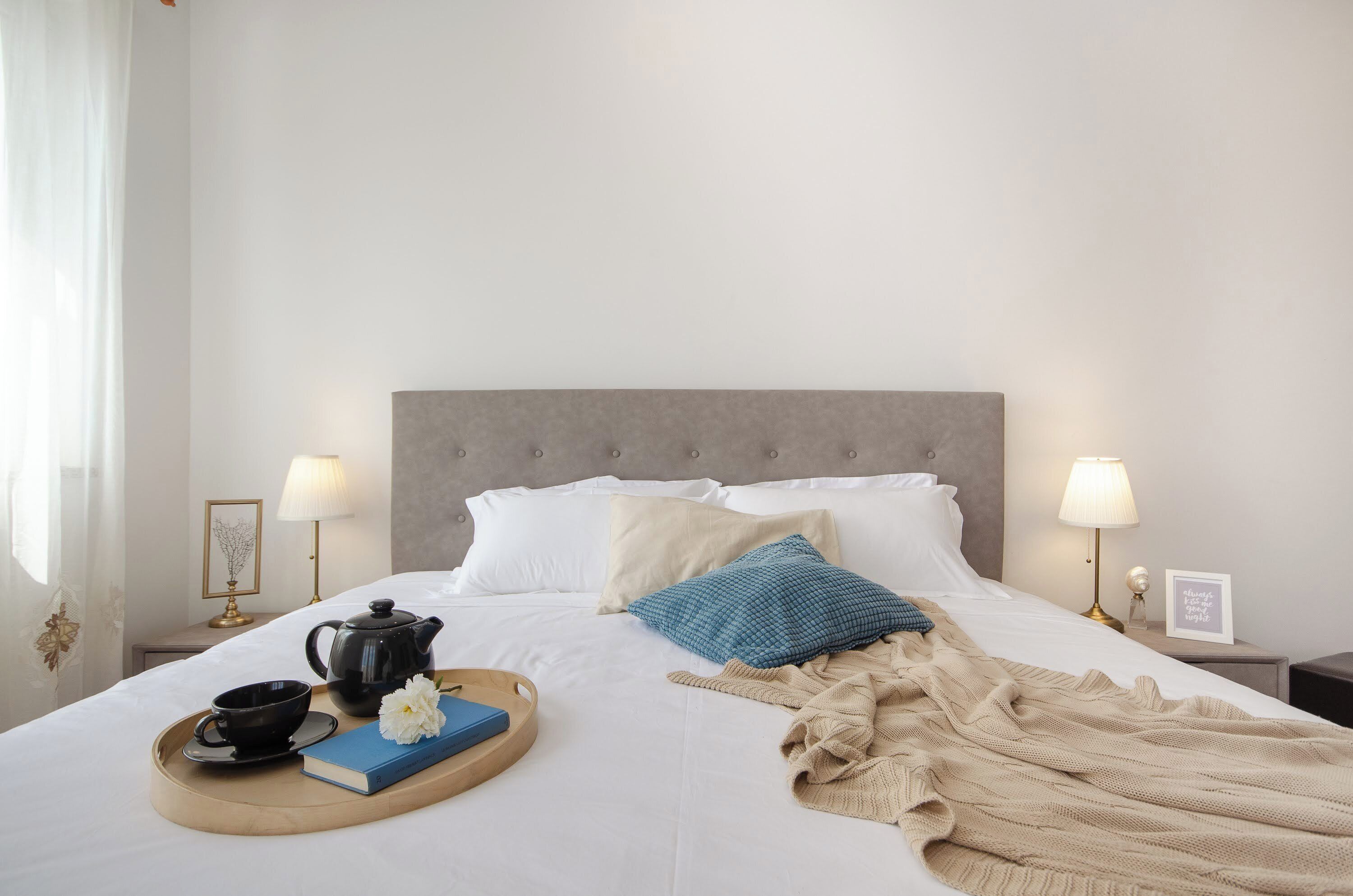 A neatly made bed with pillows, a bedside table with a lamp, a tray with a teapot, a book, and a flower.