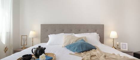 A neatly made bed with pillows, a bedside table with a lamp, a tray with a teapot, a book, and a flower.