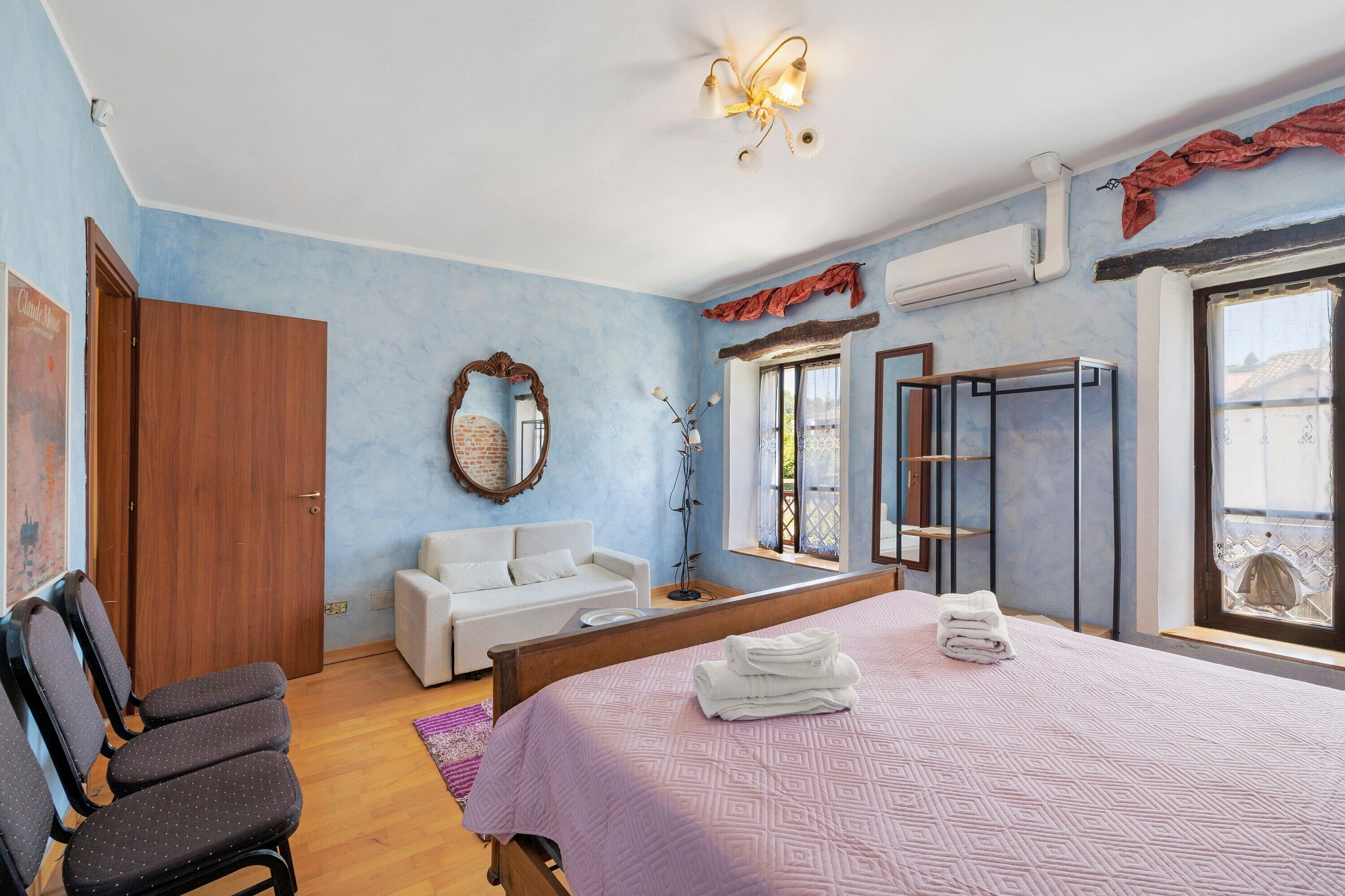A bedroom with a bed, a white sofa, a wooden wardrobe, a mirror, and a window with a view.