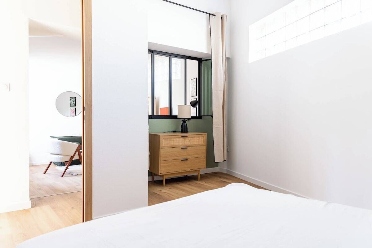 Une chambre à coucher avec un lit, une commode, une lampe et une fenêtre avec des rideaux.