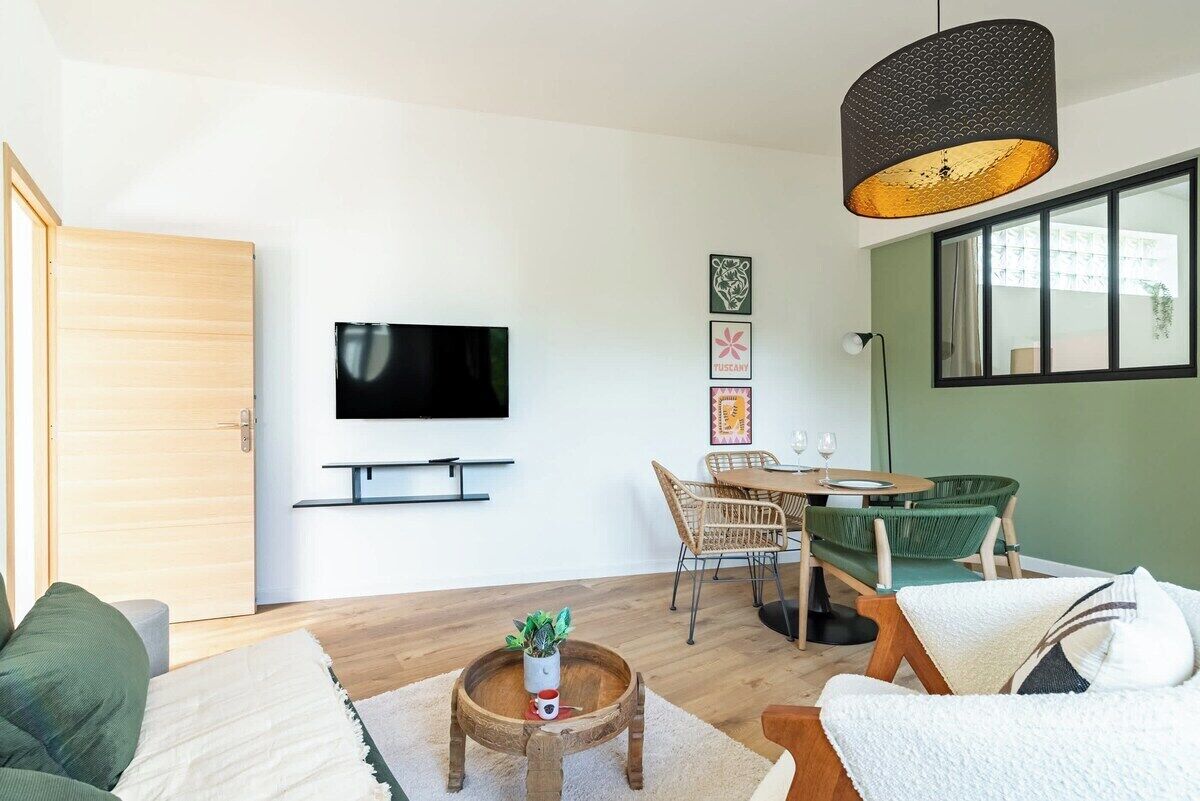 Un salon moderne doté d’un téléviseur à écran plat, de meubles en osier, d’une table basse ronde en bois et d’une grande fenêtre.