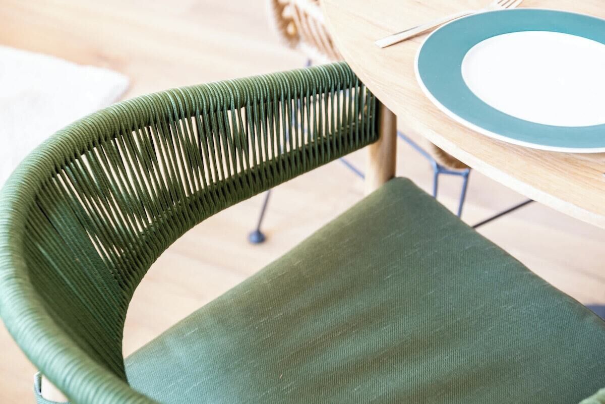 A green woven chair with a cushioned seat and backrest, positioned at a wooden table with a blue rimmed plate and a fork.