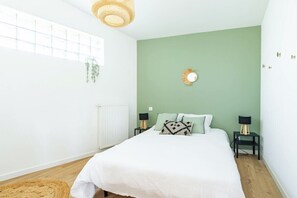 A bedroom with a bed, two bedside tables, a radiator, a window, and a hanging plant.