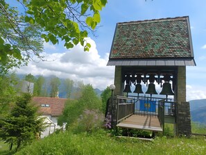 Un clocher doté d’une plateforme en bois et d’une rambarde en métal, entouré de verdure et avec une chaîne de montagnes en arrière-plan.