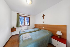 Une chambre avec deux lits, une tête de lit en bois, une table de chevet avec une lampe, une fenêtre avec des rideaux et un crucifix fixé au mur.