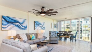 A spacious living room with a gray sectional sofa, a coffee table, and a ceiling fan. There is a dining area with a table and chairs, and large windows offering a view of the outside. - Captains Quarters 318: charming, steps to ocean and pool (Saint Augustine)