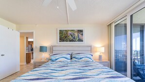 A bedroom with a large bed, two bedside lamps, a ceiling fan, a painting above the bed, and a sliding glass door leading to a balcony. - Captains Quarters 318: charming, steps to ocean and pool (Saint Augustine)