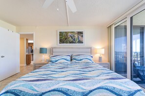 A bedroom with a large bed, two bedside lamps, a ceiling fan, a painting above the bed, and a sliding glass door leading to a balcony. - Captains Quarters 318: charming, steps to ocean and pool (Saint Augustine)