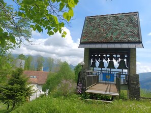 Un clocher doté d’une plateforme en bois et d’une porte bleue, entouré de verdure et de montagnes.