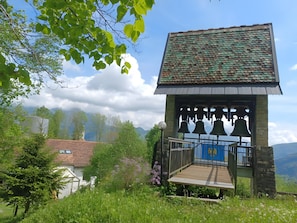 Un clocher doté d’une plateforme en bois et d’une porte bleue, entouré de verdure et de montagnes.