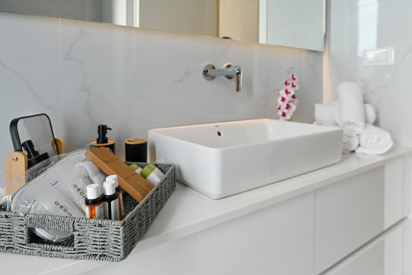 A modern bathroom with a white sink, a mirror, and toiletries in a basket.