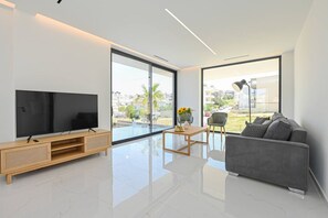 Ein modernes Wohnzimmer mit einem Flachbildfernseher, einem grauen Sofa, einer Holz-Unterhaltungseinheit und großen Fenstern mit Blick ins Freie.