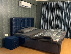 Une chambre à coucher comprenant un lit, une table de chevet, une commode, un climatiseur fixé au mur et une fenêtre avec des rideaux.
