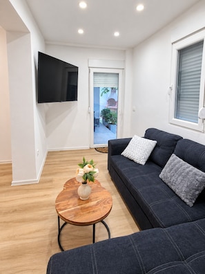 A modern living room with a dark sofa, a round wooden coffee table with a vase of flowers, a flat-screen TV mounted on the wall, and a window with blinds. - Apartment Perla (Vir)
