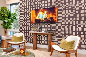 Een woonkamer met een televisie waarop een haard te zien is, twee fauteuils, een bijzettafeltje met een plant en boeken.