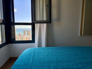 Ein Zimmer mit einem Bett, einem Fenster mit Vorhängen und Blick auf Gebäude und das Meer.