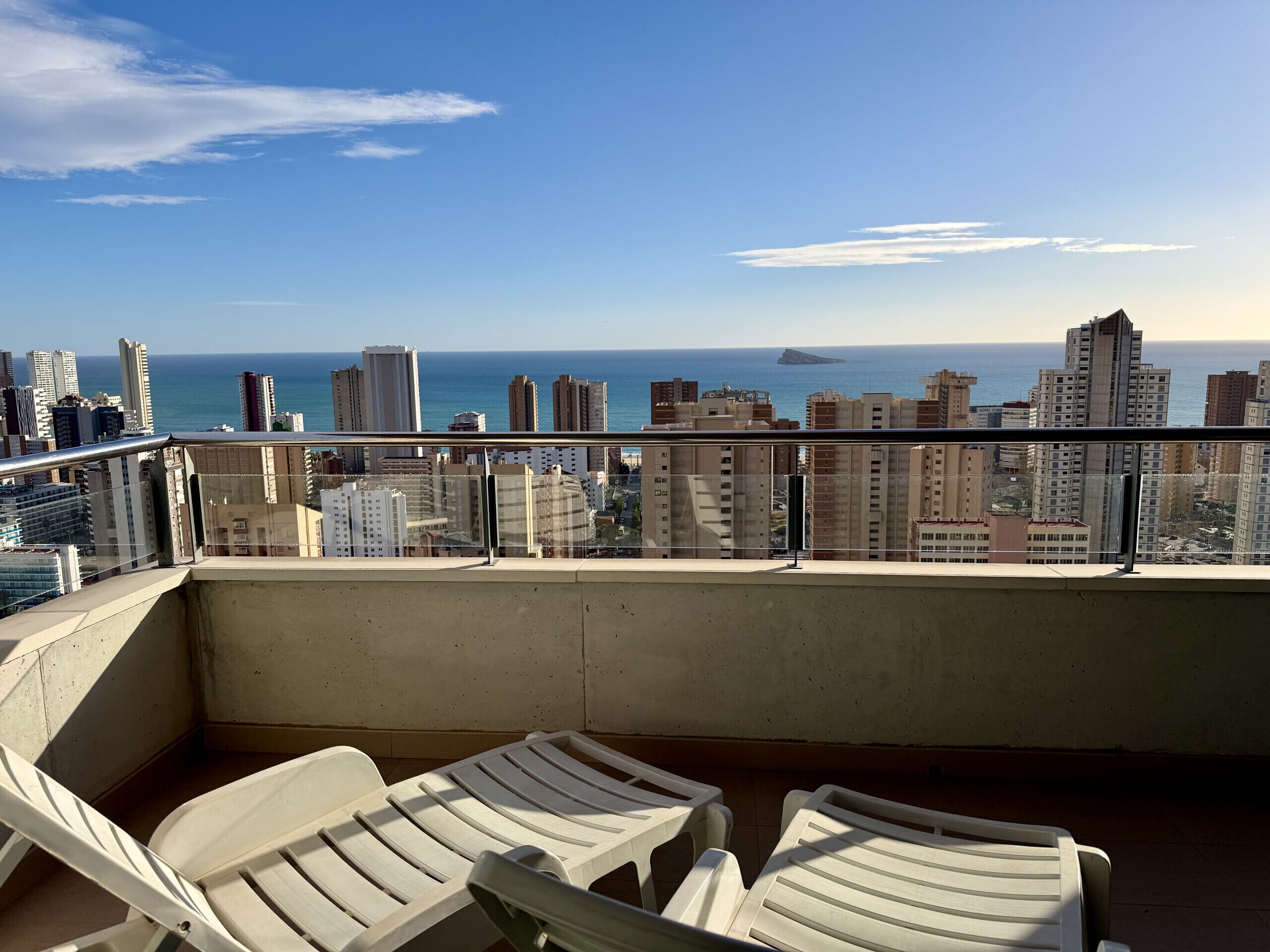 A balcony with two white plastic chairs, a view of a cityscape with tall buildings and a body of water in the background.