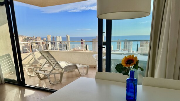 Un balcon avec une table blanche, un vase bleu contenant un tournesol, et une vue sur la ville et la mer.