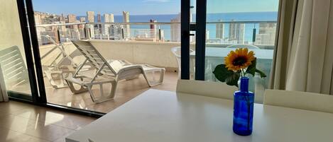 Un balcon avec une table blanche, un vase bleu contenant un tournesol, et une vue sur la ville et la mer.