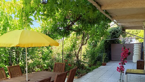 Een terras buiten met een houten tafel en stoelen, een gele parasol en een betegelde vloer.