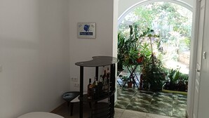 Een kamer met een witte tafel, een zwart schap met flessen, een bord aan de muur, en uitzicht op een tuin door een boogvormige deuropening.