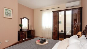 A bedroom with a bed, bedside table, dresser, mirror, and a window with sheer curtains.