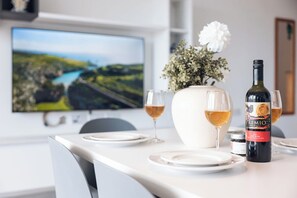 Una mesa de comedor dispuesta con platos, vasos, una botella de vino y un florero con flores. En la pared detrás de la mesa hay un televisor que muestra un paisaje pintoresco.