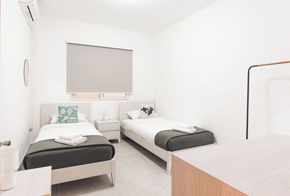 A modern hotel room with two single beds, a small bedside table with a lamp, a large window with a blind, and a wooden desk.