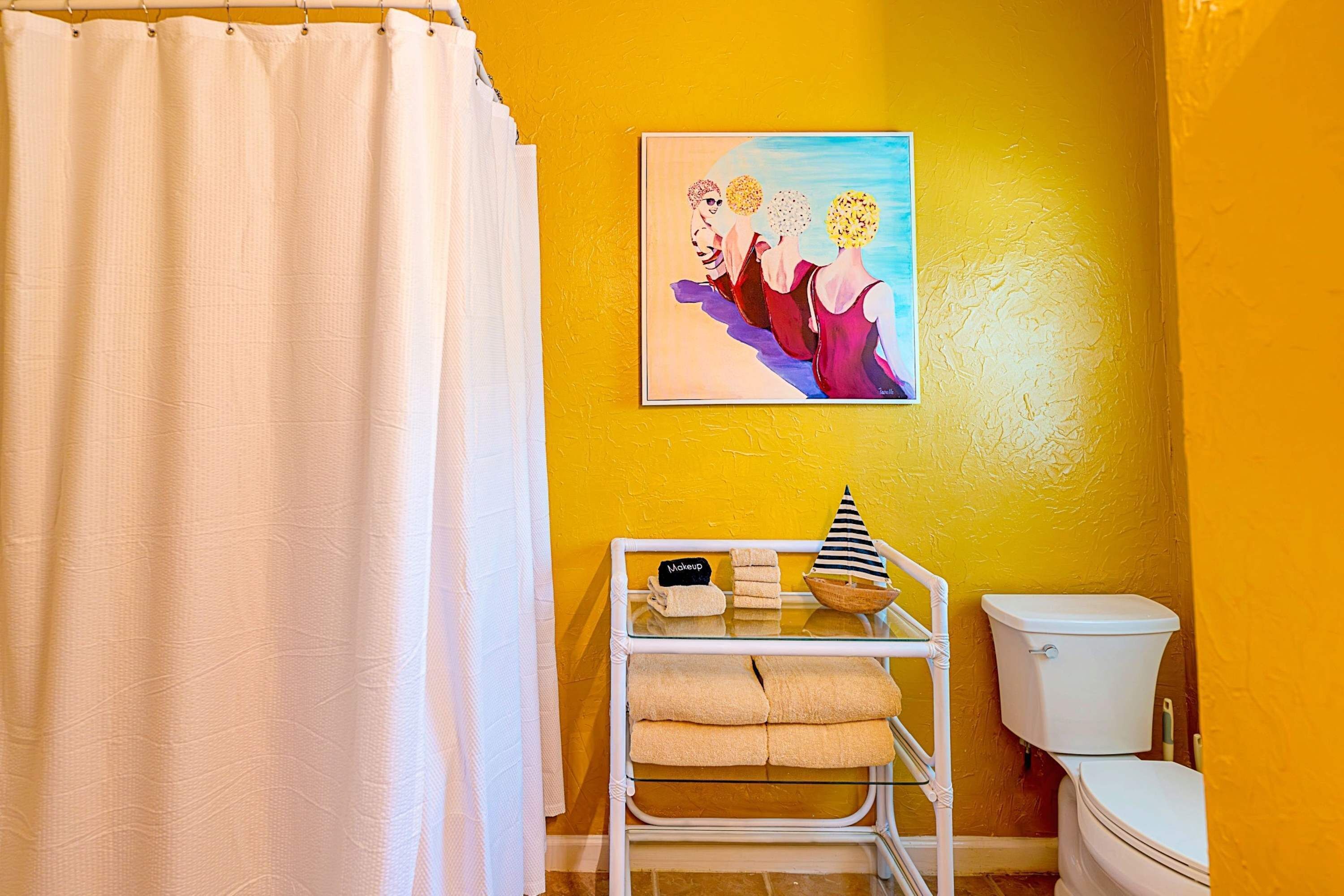 Un baño con una pared amarilla, una cortina de ducha blanca, un inodoro blanco y un perchero con toallas y un gorro.