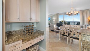 A kitchen with granite countertops, wooden cabinets, and a wine rack. There is a dining area with a table and chairs, and a view of the ocean through large windows. - Indigo West 304 by Liquid Life (Pensacola)