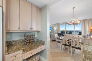 A kitchen with granite countertops, wooden cabinets, and a wine rack. There is a dining area with a table and chairs, and a view of the ocean through large windows. - Indigo West 304 by Liquid Life (Pensacola)