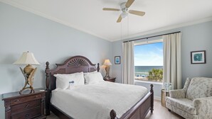 A bedroom with a large bed, a ceiling fan, a nightstand, a chair, and a window with a view of the beach. - Indigo West 304 by Liquid Life (Pensacola)