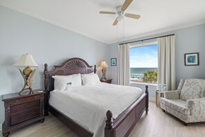 A bedroom with a large bed, a ceiling fan, a nightstand, a chair, and a window with a view of the beach. - Indigo West 304 by Liquid Life (Pensacola)