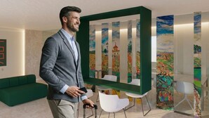 A man in a grey suit holding a phone, standing in a modern room with a green sofa, white chairs, and colorful abstract paintings on the walls. - Hotel Ogar Tomelloso (Tomelloso)