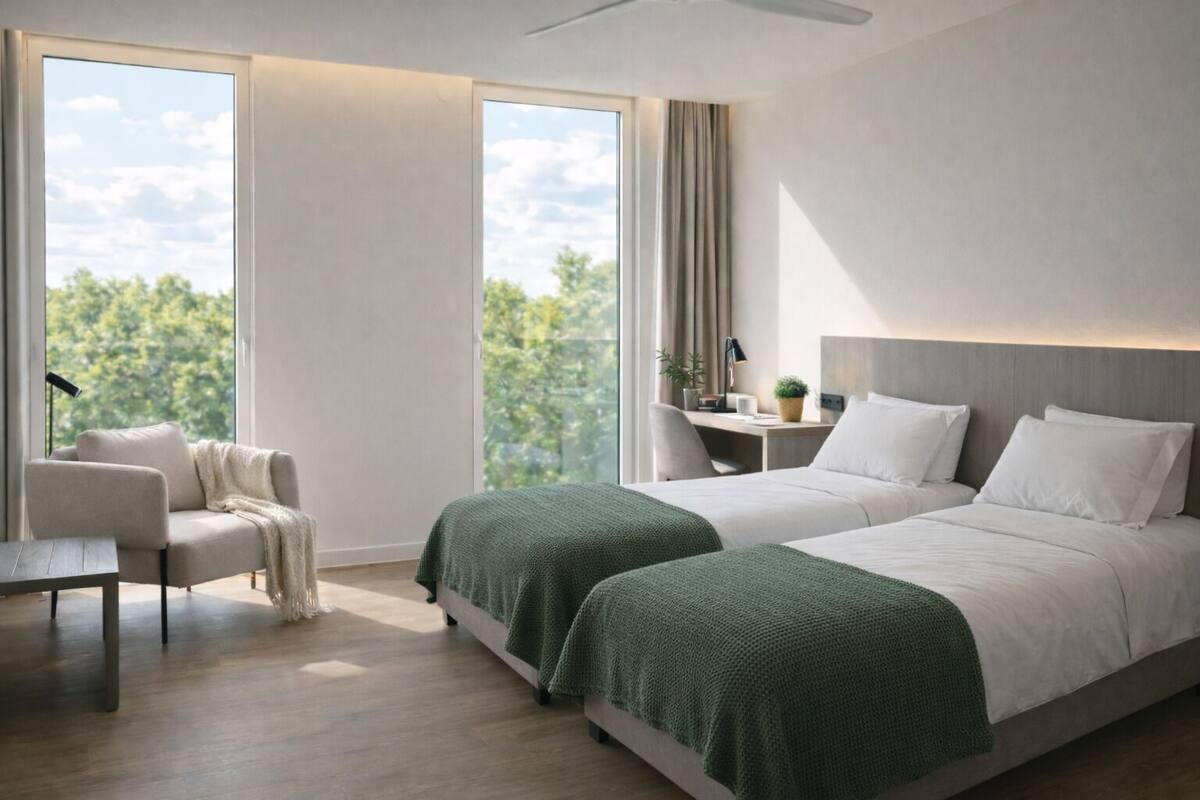 A modern hotel room with two beds, a desk, a chair, and large windows with a view of trees.