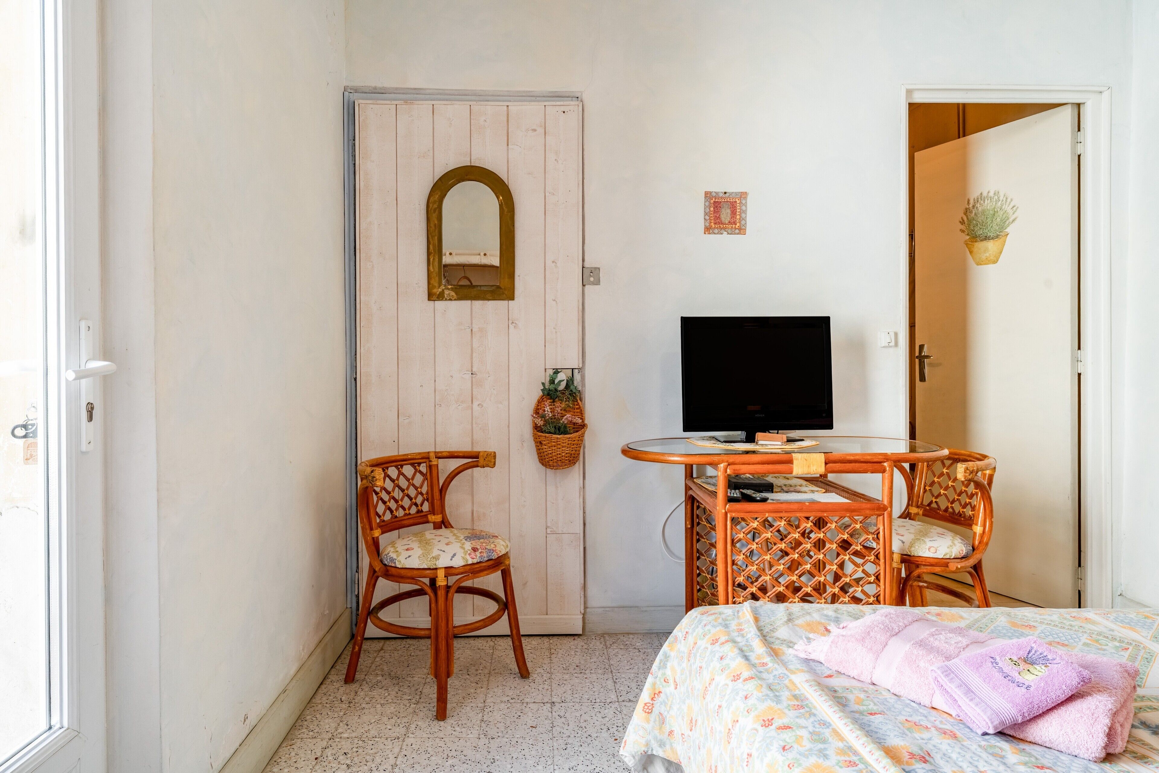 Habitació amb un llit, una televisió, dues cadires de vimet, una taula petita, una porta amb un mirall i una planta penjant a la paret.