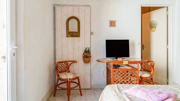 Habitació amb un llit, una televisió, dues cadires de vimet, una taula petita, una porta amb un mirall i una planta penjant a la paret.