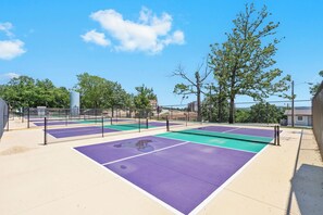 An outdoor tennis court with multiple courts, surrounded by trees and buildings.