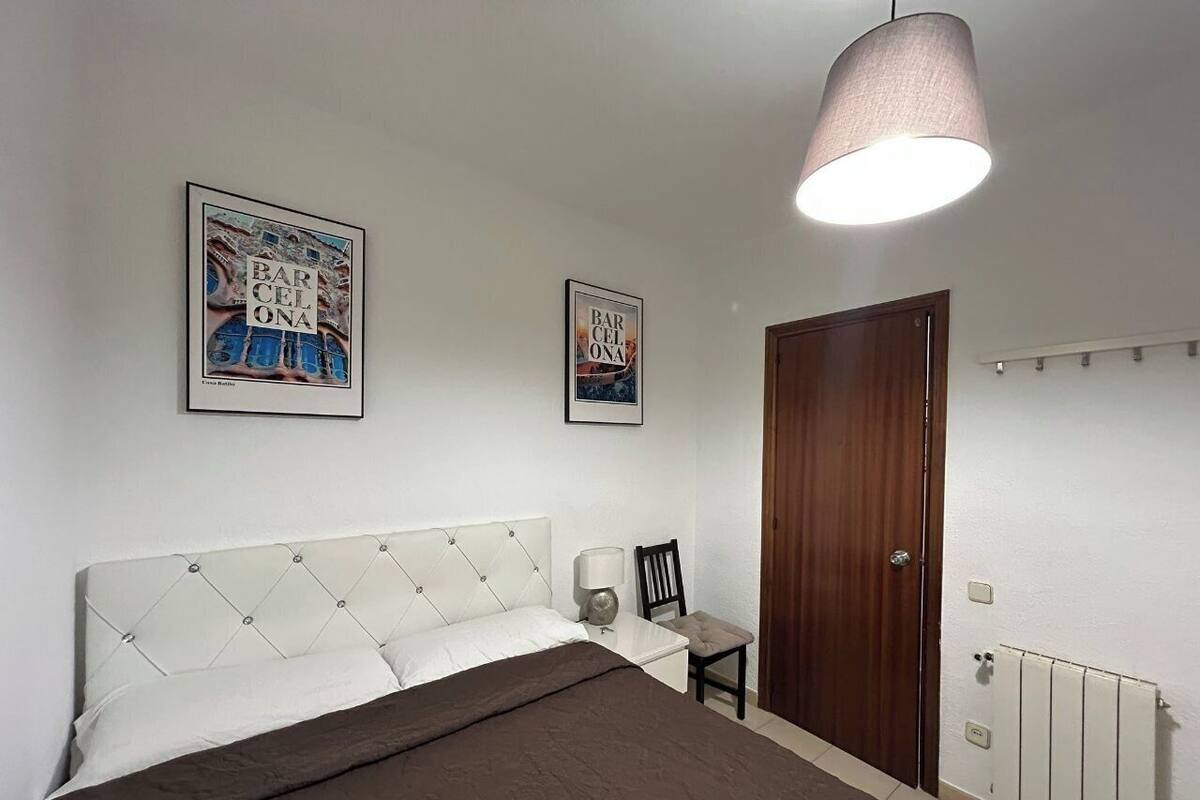 Un dormitorio con una cama, una silla, una mesita de noche, una lámpara, una puerta y dos cuadros en la pared.